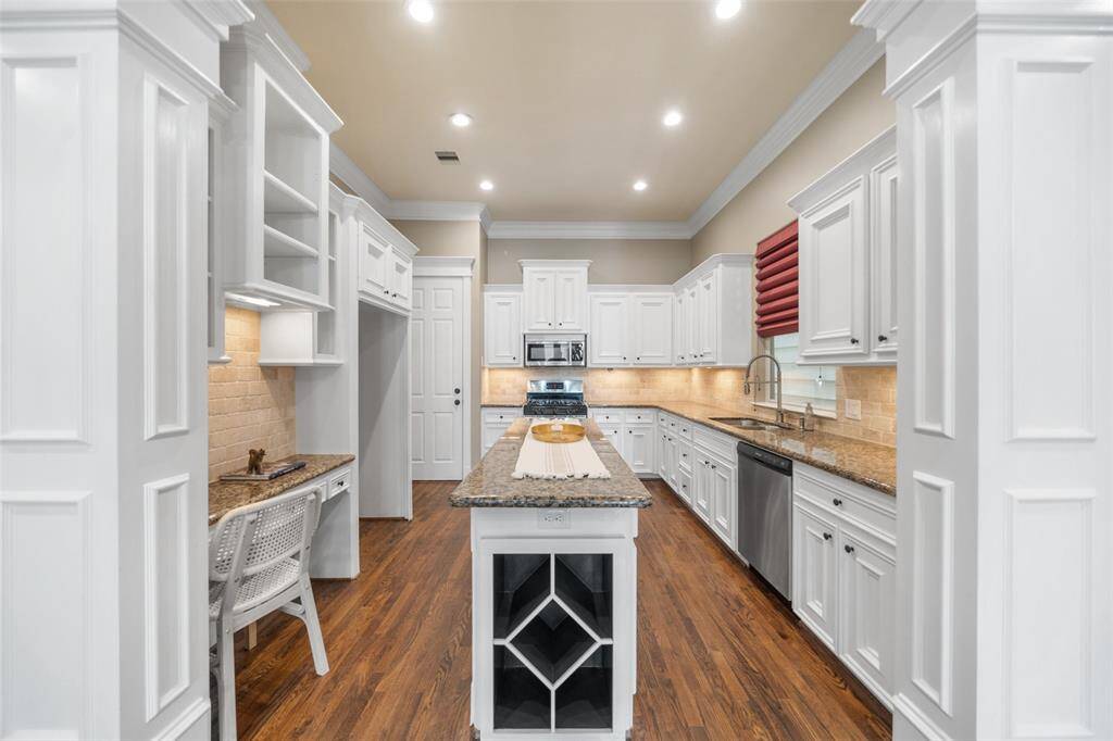 This spacious kitchen features elegant white cabinetry, granite countertops, and a central island with a built-in wine rack. The warm wood flooring and recessed lighting create a cozy yet modern atmosphere, complemented by stainless steel appliances and a convenient desk area.