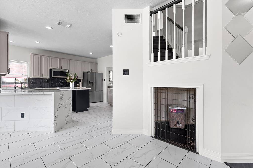 This photo showcases a modern kitchen and entryway area in a home. The kitchen features sleek cabinetry, stainless steel appliances, and a granite countertops & island. The space is open and bright with white tile flooring and recessed lighting. The staircase has a classic railing, and there’s a small nook underneath.