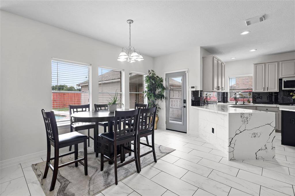 This bright dining area features a modern table set on a sleek tile floor. Large windows offer ample natural light, complementing the adjacent contemporary kitchen with marble countertops and stylish cabinetry. The space is open and inviting, ideal for entertaining.