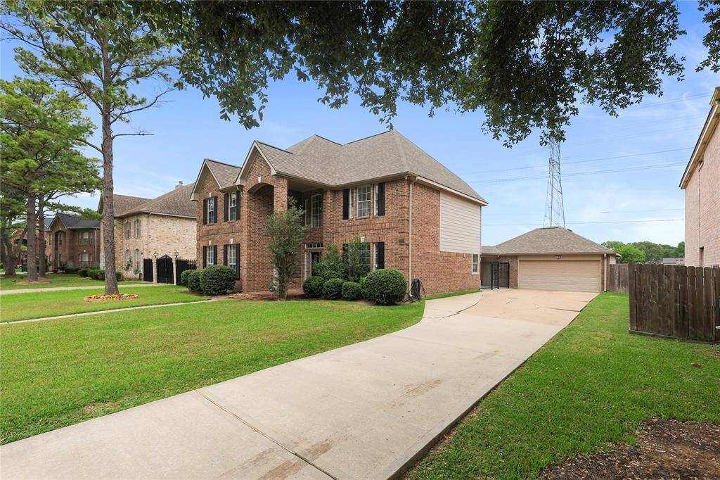 This photo features a spacious two-story brick home with a well-maintained lawn and landscaping. It has a long driveway leading to a detached garage. Mature trees lining the street.