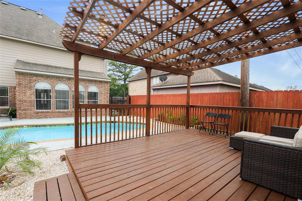Backyard with a spacious wooden deck featuring a pergola for shade. The deck overlooks a well-maintained pool, bordered by a brick house, fenced backyard creating a private and relaxing outdoor space.