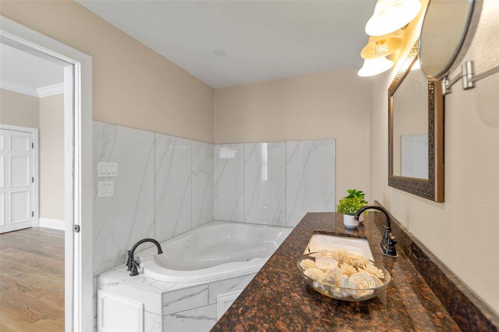 This modern primary bathroom features a built-in corner bathtub with white marble-like tiling, a granite countertop with a sink.