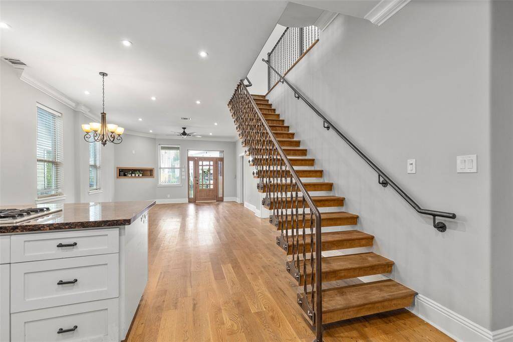 View from the kitchen to the front door that shows a bright, open-concept space with hardwood floors, featuring a modern kitchen with a large island, elegant lighting, and a view into a dining area. A graceful floating staircase with wooden steps and a wrought-iron baluster leads to the upper level, enhancing the home's spacious and upscale feel.