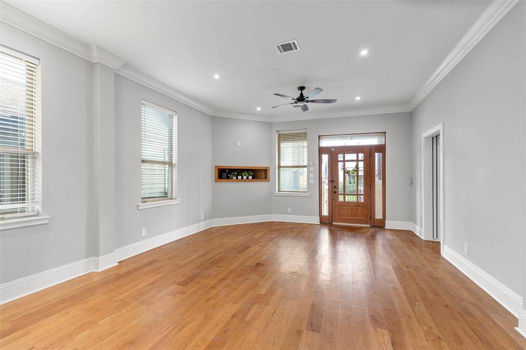This is a spacious, well-lit room with polished hardwood floors, elegant crown molding, and multiple windows for natural light, a built-in shelf for electronic equipment.