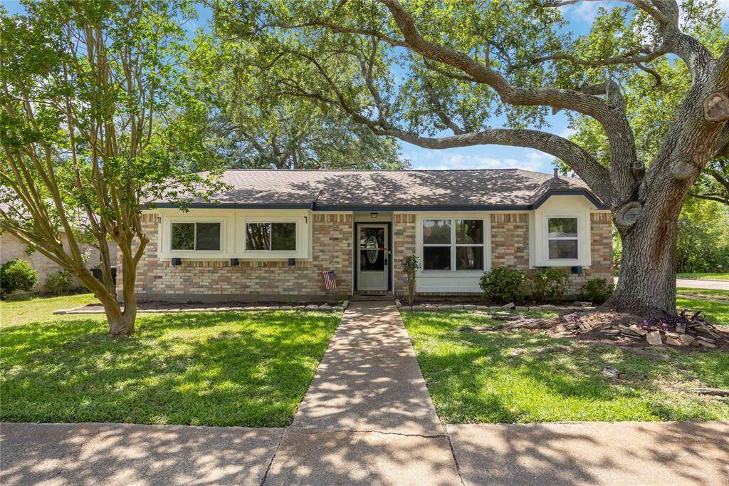 This charming single-story home features a brick exterior with a welcoming front walkway. It's surrounded by lush greenery and mature trees, offering a serene and inviting atmosphere.