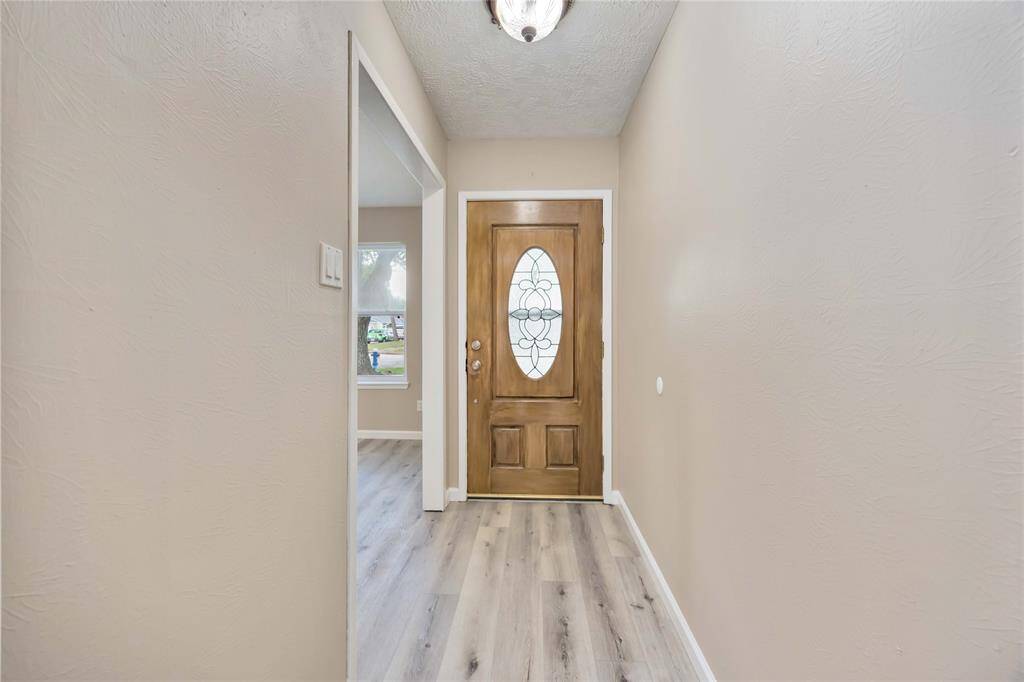 This photo shows a welcoming entryway with a wooden front door featuring an oval glass design. The hallway has light-colored walls and wood-style flooring, creating a bright and inviting atmosphere. A glimpse of a room with a window is visible to the left.