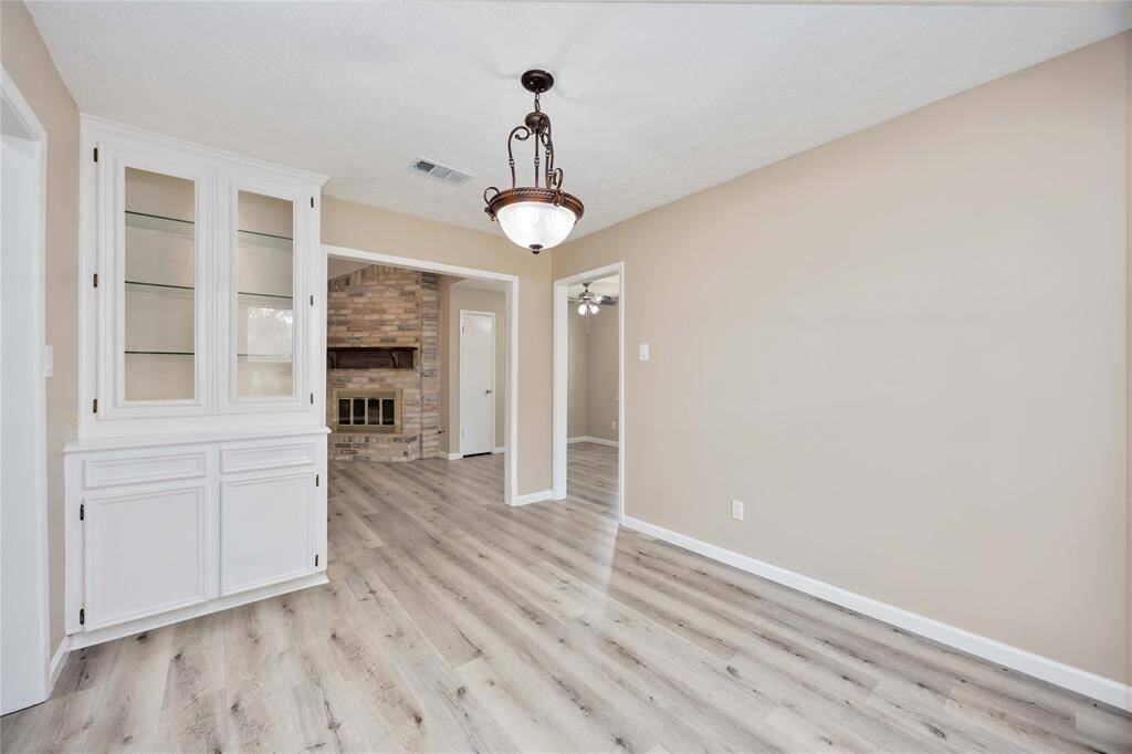 This room features light wood flooring and neutral walls, creating a bright, welcoming space. There's a built-in white cabinet with glass shelves for display, and a chandelier adds elegance. An open doorway leads to a cozy area with a brick fireplace.