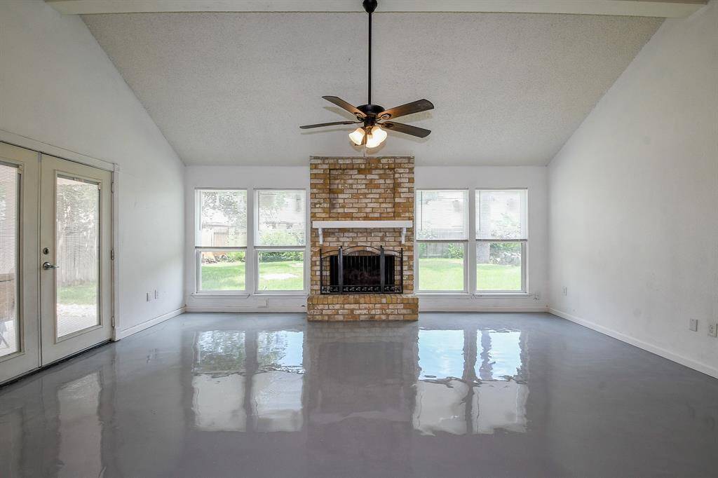 This spacious living room features a high ceiling, a central brick fireplace, and large windows offering ample natural light. The glossy floor adds a modern touch, while the double doors open to the outdoors, enhancing the room's airy feel.