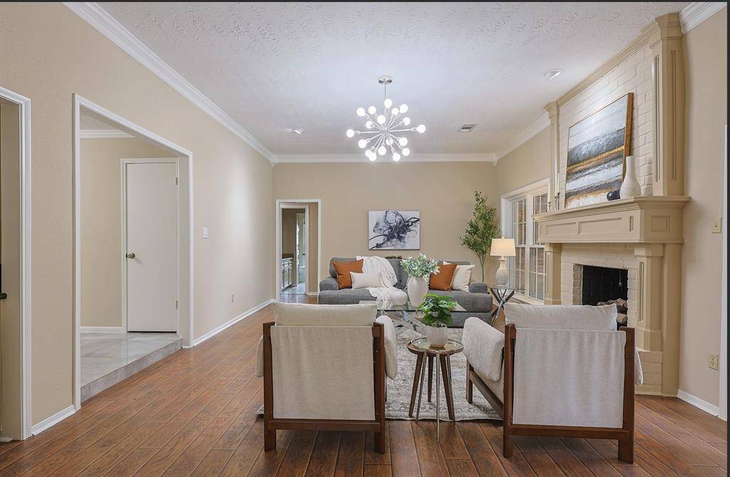 This spacious living room features a stylish chandelier, and a cozy brick fireplace. Large windows and French doors provide ample natural light, while the open layout flows into an adjacent room, creating a welcoming atmosphere.