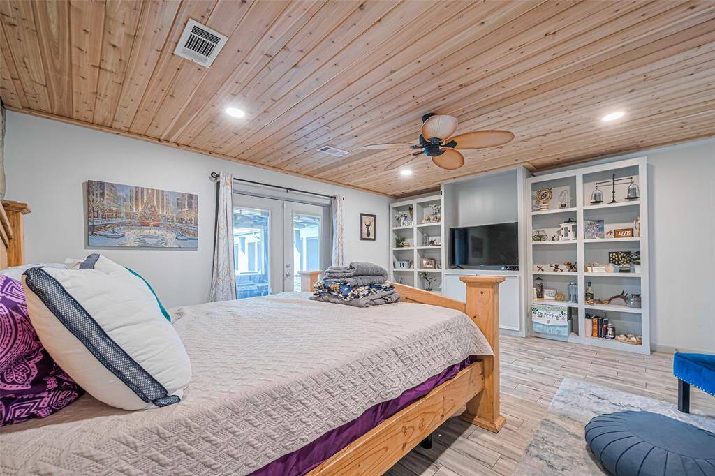 This cozy primary bedroom features a wooden ceiling with recessed lighting and a ceiling fan. It includes built-in shelving for storage, and large glass doors leading outside.