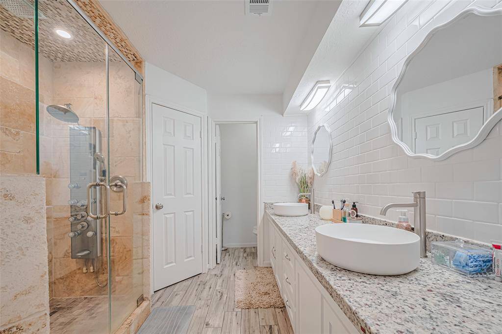 This bathroom offers a modern design with a glass-enclosed shower, dual sinks on a granite countertop, and white subway-tile walls. The wood-look flooring adds warmth and elegance.