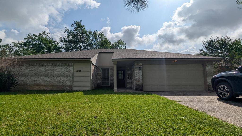 This photo shows a single-story house with a two-car garage, light brick exterior, and a well-maintained front lawn. The property is surrounded by mature trees, providing a pleasant and shaded environment.