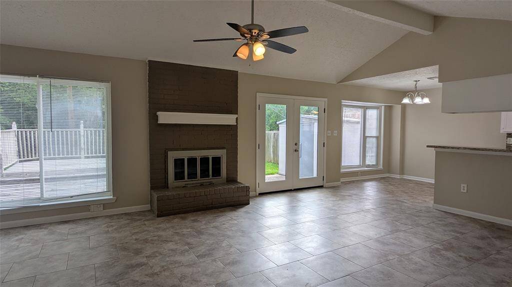 This spacious living area features a modern tiled floor, a cozy brick fireplace, and ample natural light from large windows. The room includes a ceiling fan and opens to a patio through French doors, with an adjacent dining area and open kitchen concept.