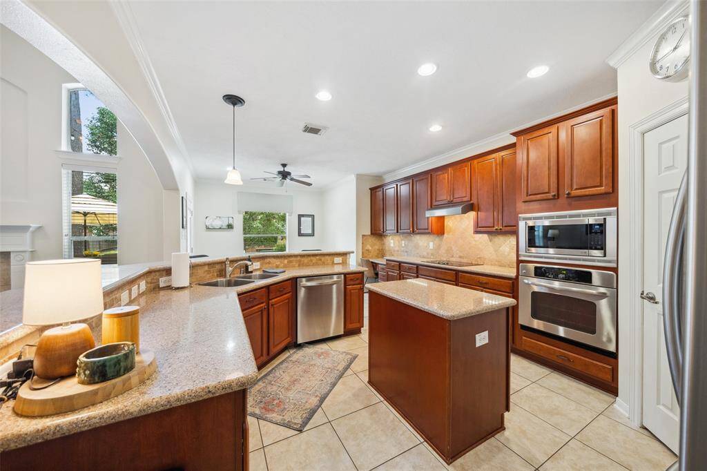 Spacious kitchen featuring granite countertops, stainless steel appliances, and rich wood cabinetry. Includes a central island, tile flooring, and an open layout with large windows for natural light.
