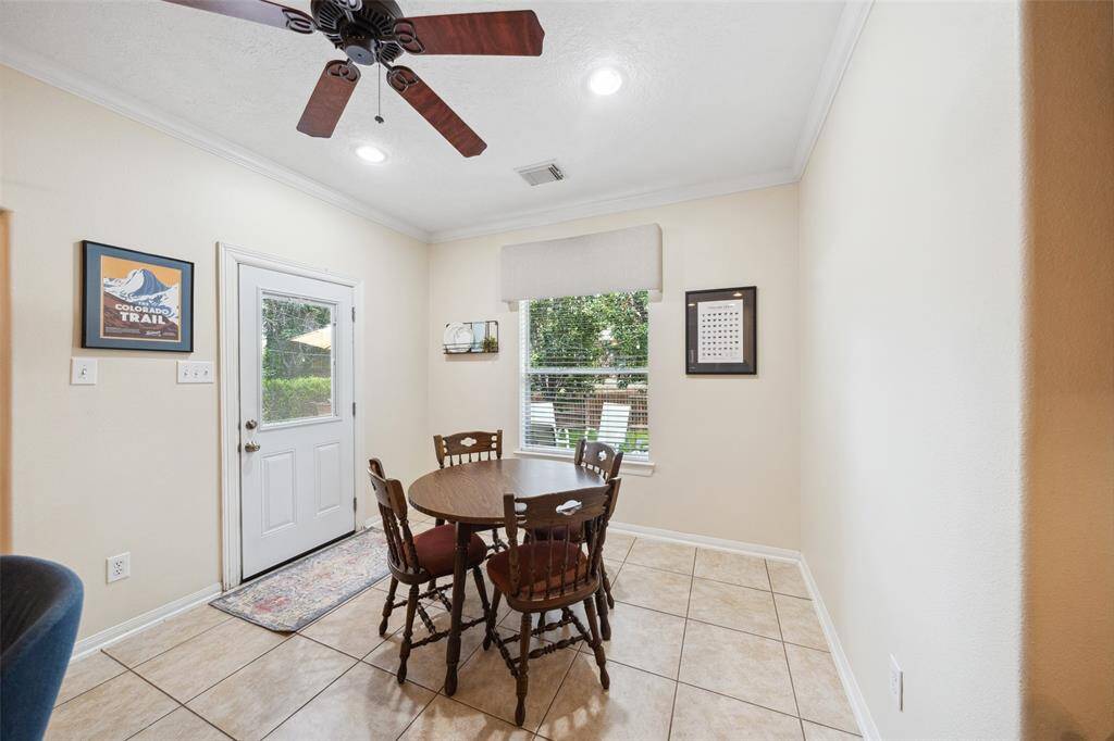 Dining area featuring large tiles, neutral walls, and a ceiling fan. Natural light streams in through a window and a glass-paneled door, offering a view of a lush backyard.