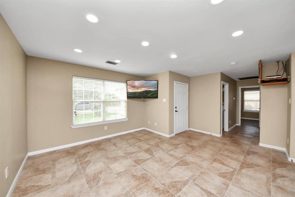 A spacious, open-concept living area with tiled flooring and neutral-toned walls. The kitchen features modern cabinetry and appliances, with ample lighting from recessed ceiling lights. A door at the back leads to the outside, offering easy access to outdoor spaces.