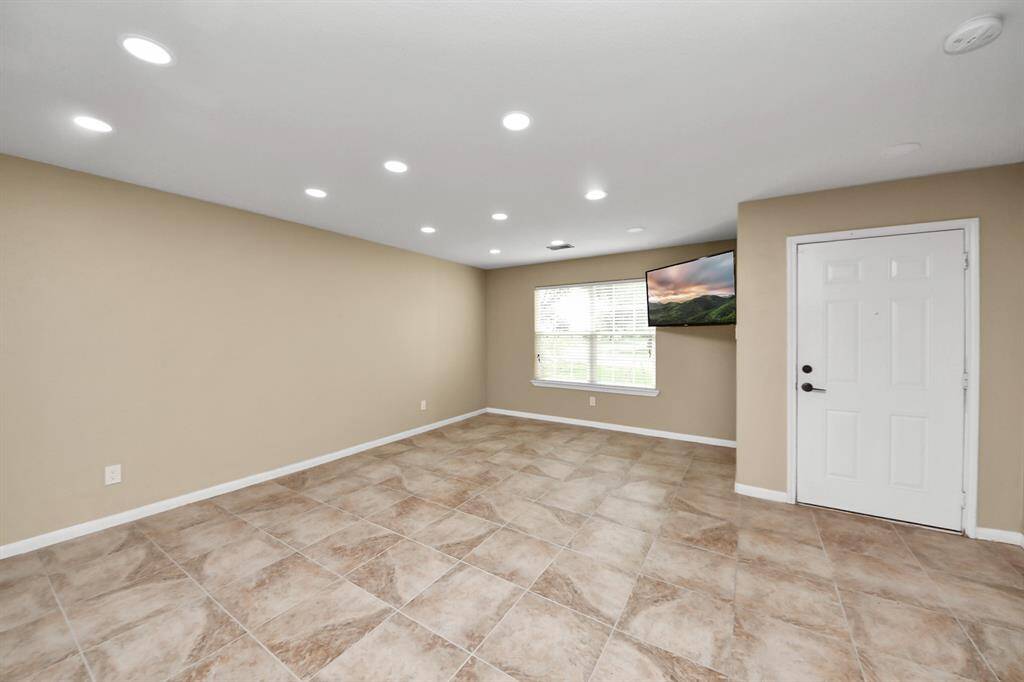Spacious living area with neutral tones, featuring tiled floors, recessed lighting, and a large window for natural light. Includes a wall-mounted TV and a clean, modern entry door.