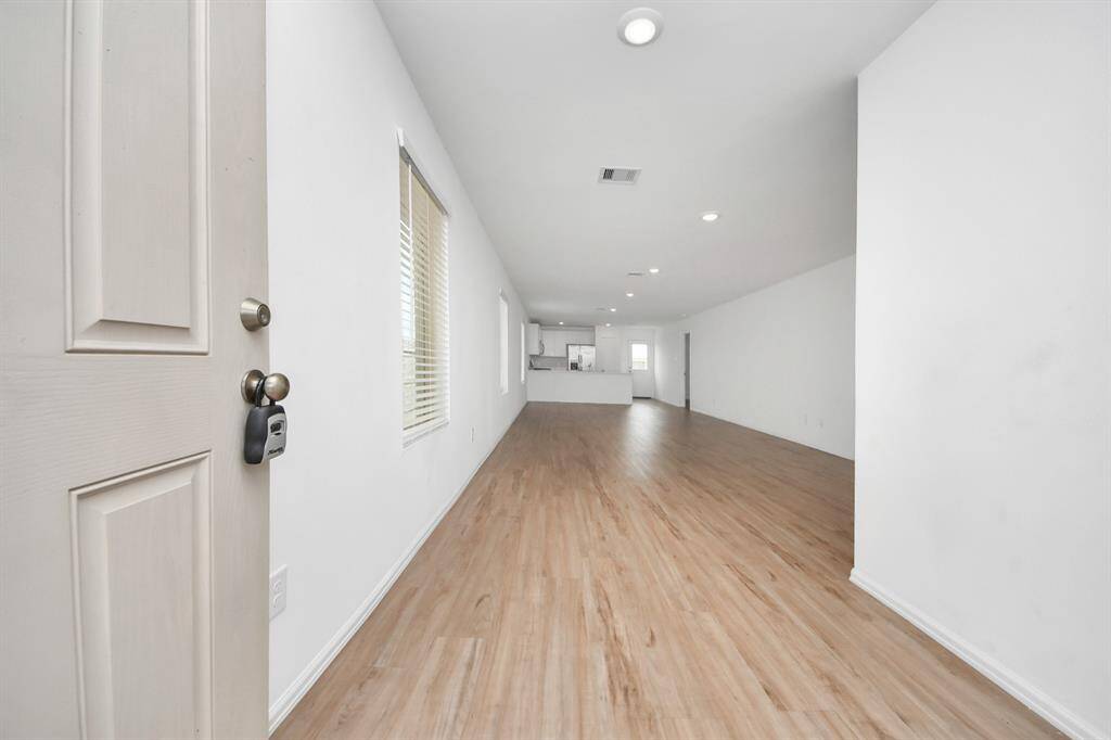 A spacious, open-concept living area with light wood flooring and white walls, leading to a modern kitchen. Recessed lighting provides a bright, airy feel, and a window allows for natural light.
