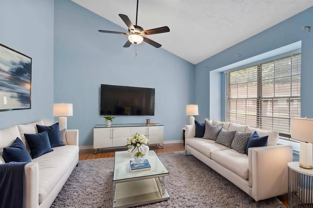 This inviting living room features soaring vaulted ceilings, a large picture window that fills the space with natural light, and soft blue walls that create a calming atmosphere. The open layout offers plenty of space for gathering or relaxing, while the ceiling fan adds comfort and functionality. With beautiful flooring and a neutral color palette, this space is ready to be customized to fit your style.