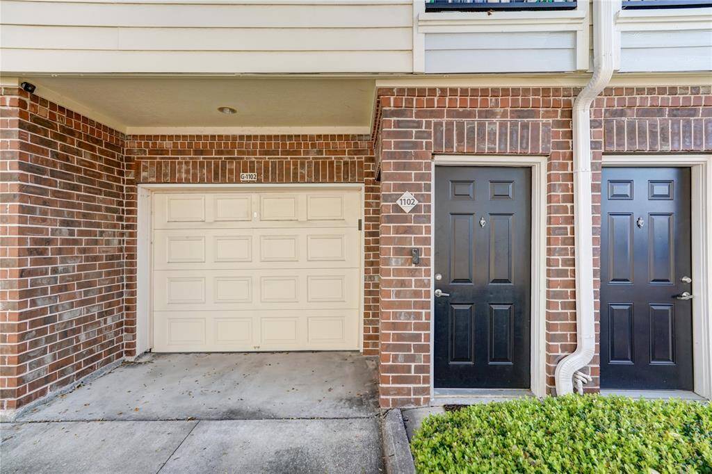 This photo shows the front of a townhouse featuring a single-car garage and two black entry doors, all framed with red brick. There's a small garden area with greenery beside the doors.
