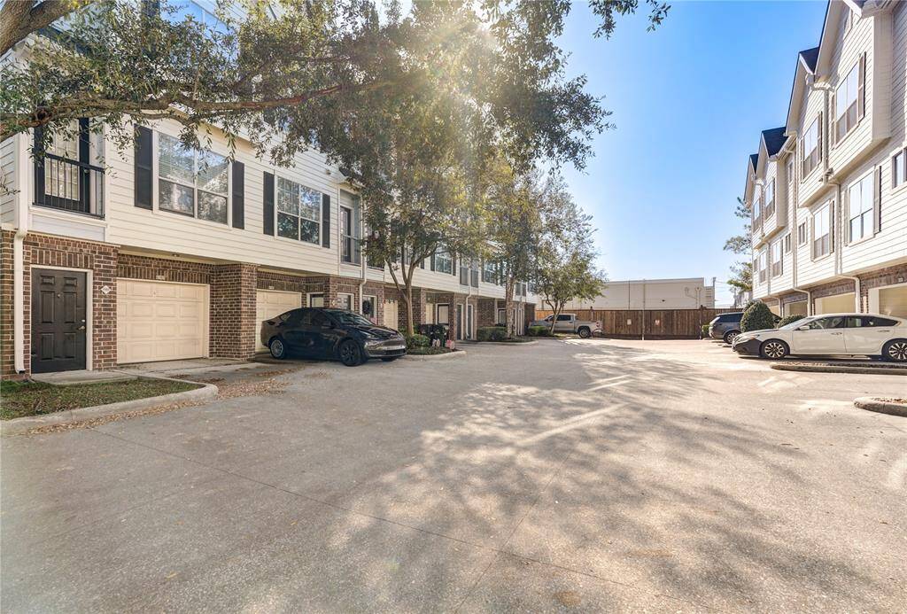 This photo shows a residential complex with multi-story townhouses featuring brick and siding exteriors. The area includes garages, a driveway, and is surrounded by trees, offering a clean and inviting environment.