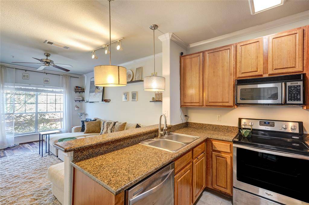 This photo showcases a cozy kitchen-living room area with warm wooden cabinets, granite countertops, and stainless steel appliances. The open layout leads to a bright living space with large windows, a ceiling fan, and a comfortable seating area.
