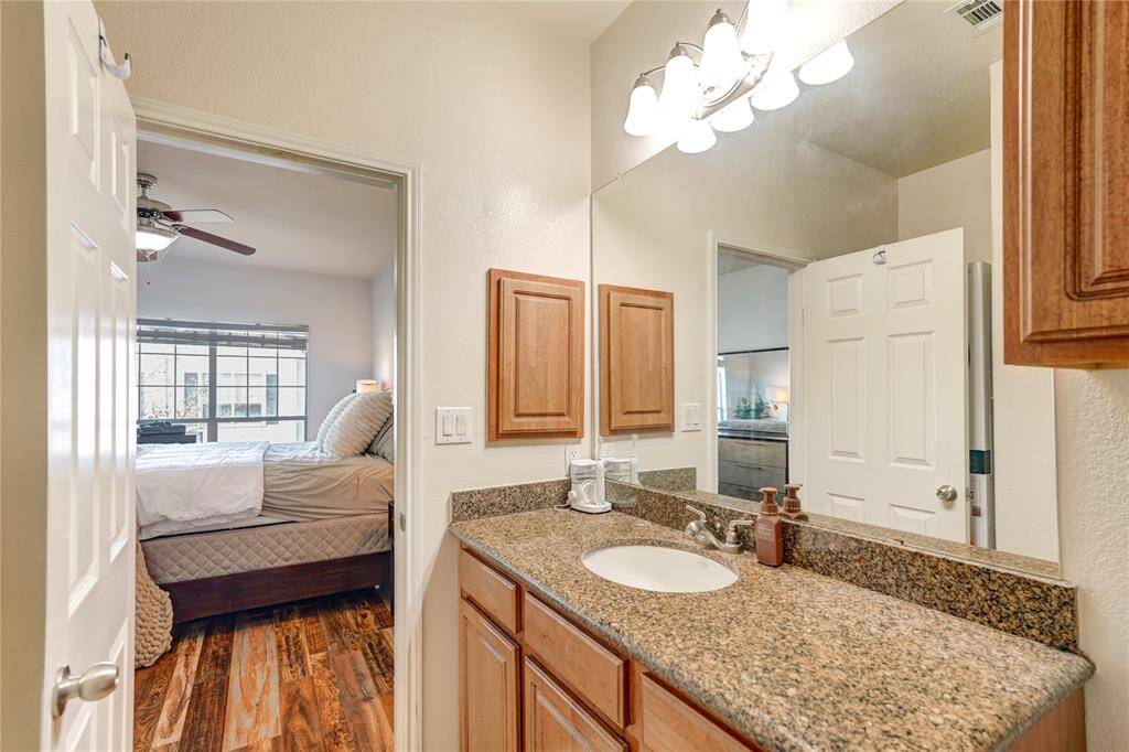 This photo shows a cozy bathroom with a granite countertop and wooden cabinetry. It features a large mirror under stylish lighting. The space opens into a bright bedroom with a ceiling fan and wood flooring, creating a warm and inviting atmosphere.