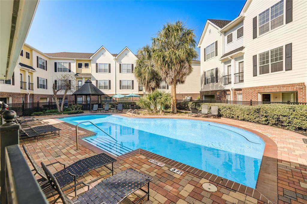 This photo showcases a well-maintained residential complex featuring a central swimming pool surrounded by a brick patio. The complex has a charming blend of brick and siding exteriors, with multiple stories and lush landscaping, including palm trees and a gazebo. Ideal for relaxing and enjoying outdoor amenities.