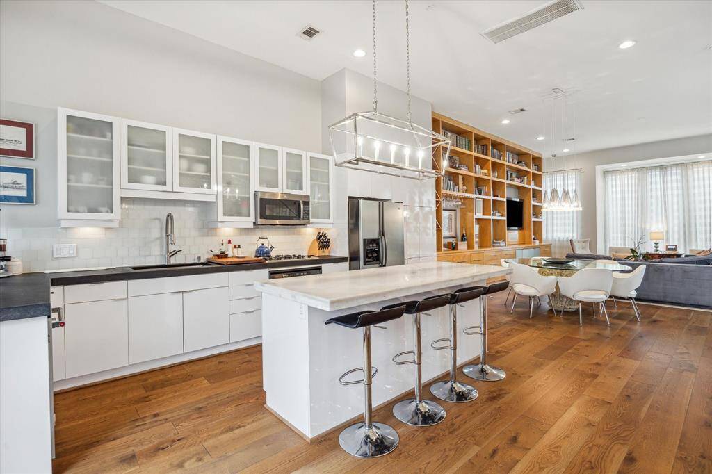 This modern open-concept kitchen and living area features sleek white cabinetry, a large island with bar seating, hardwood floors, and ample natural light. The space includes a dining area and a cozy living room with built-in shelving, creating a welcoming atmosphere for entertaining and relaxation.