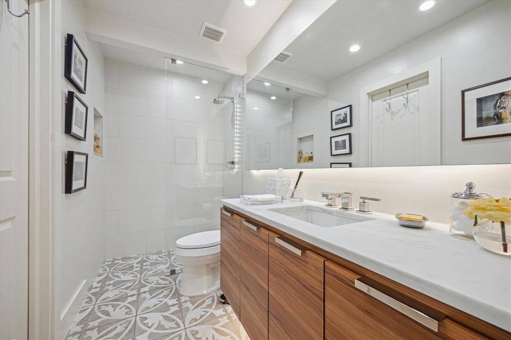 This bathroom features a modern design with a spacious glass-enclosed shower, a sleek wooden vanity with a white countertop, and stylish patterned floor tiles. The room is well-lit with recessed lighting. Can be entered from foyer or first floor bedroom.