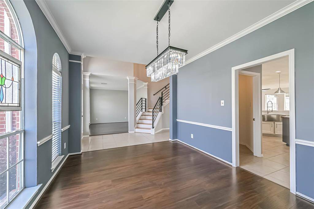 This photo showcases a modern dining area with dark wood flooring, a stylish chandelier, and large arched windows for natural light. The space opens into a kitchen with tile flooring and a view of the staircase, offering a seamless flow between rooms.