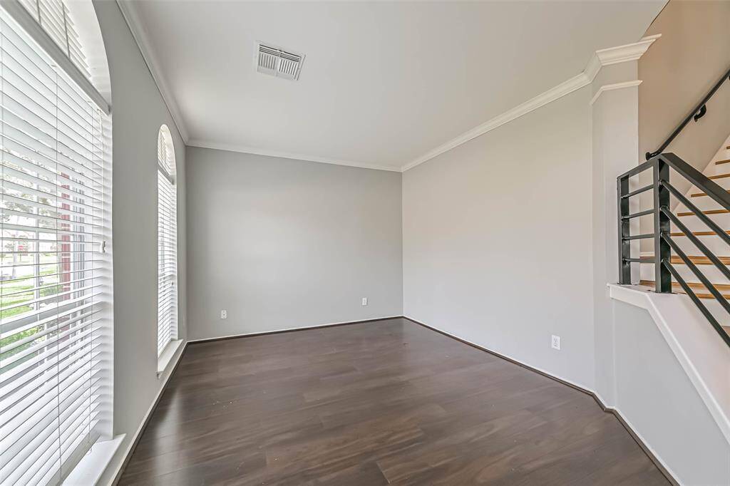 This photo shows a bright, unfurnished room with large windows, dark wood flooring, light gray walls, and a staircase with modern railing. Ideal for a living area or office space.