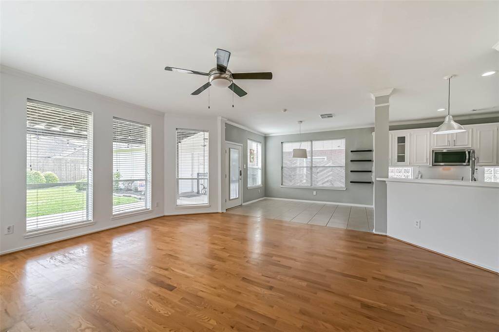 Bright, open-plan living space with hardwood floors and large windows overlooking a green backyard. Features a ceiling fan, adjacent dining area with tile flooring, and a modern kitchen with white cabinetry and stainless steel appliances.