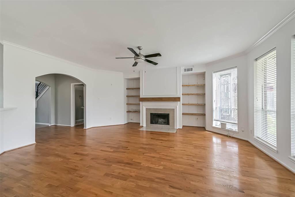 This spacious living room features hardwood floors, a cozy fireplace, built-in shelves, and large windows that let in plenty of natural light. The neutral color scheme and ceiling fan add to the room's welcoming atmosphere.