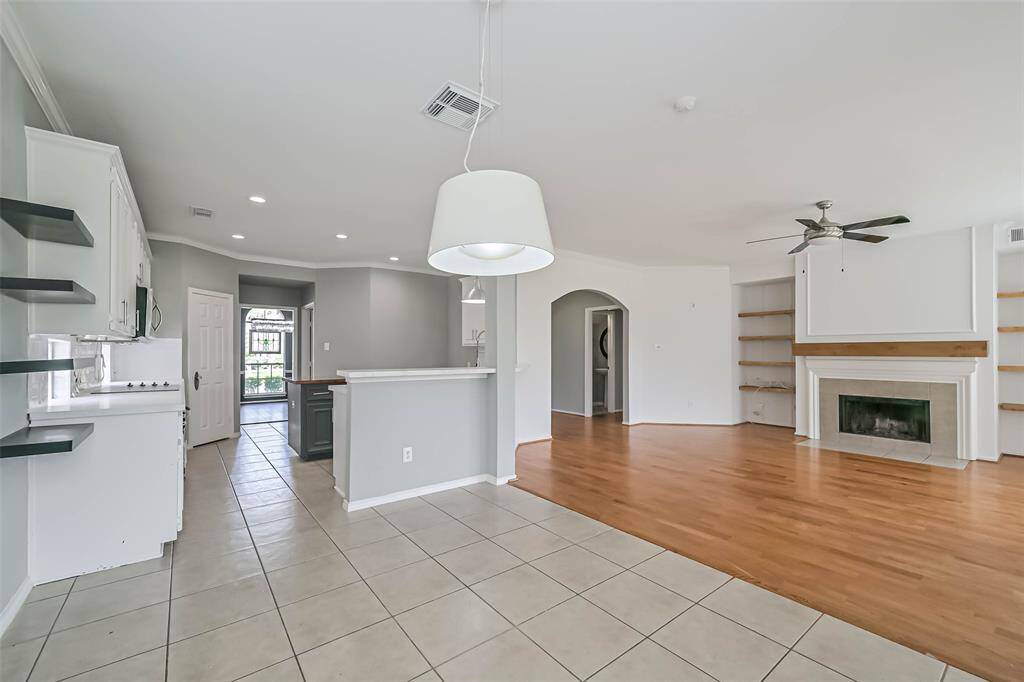 This spacious open-concept area features a kitchen with modern white cabinetry, tile flooring, and a cozy living room with wood flooring and a fireplace. Shelving surrounds the fireplace, and there's ample natural light, creating a bright and inviting atmosphere.