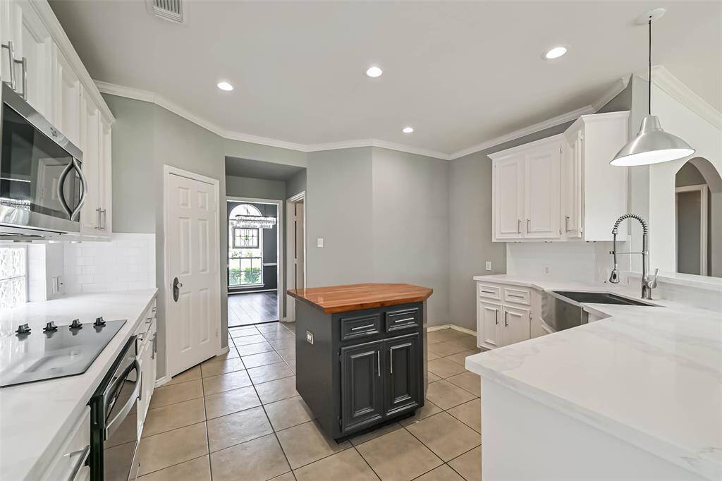 Bright kitchen with modern white cabinetry, a central island with a wooden countertop, and stainless steel appliances. Features tile flooring, recessed lighting, and a sleek faucet. Adjacent to a room with natural light streaming through a large window.