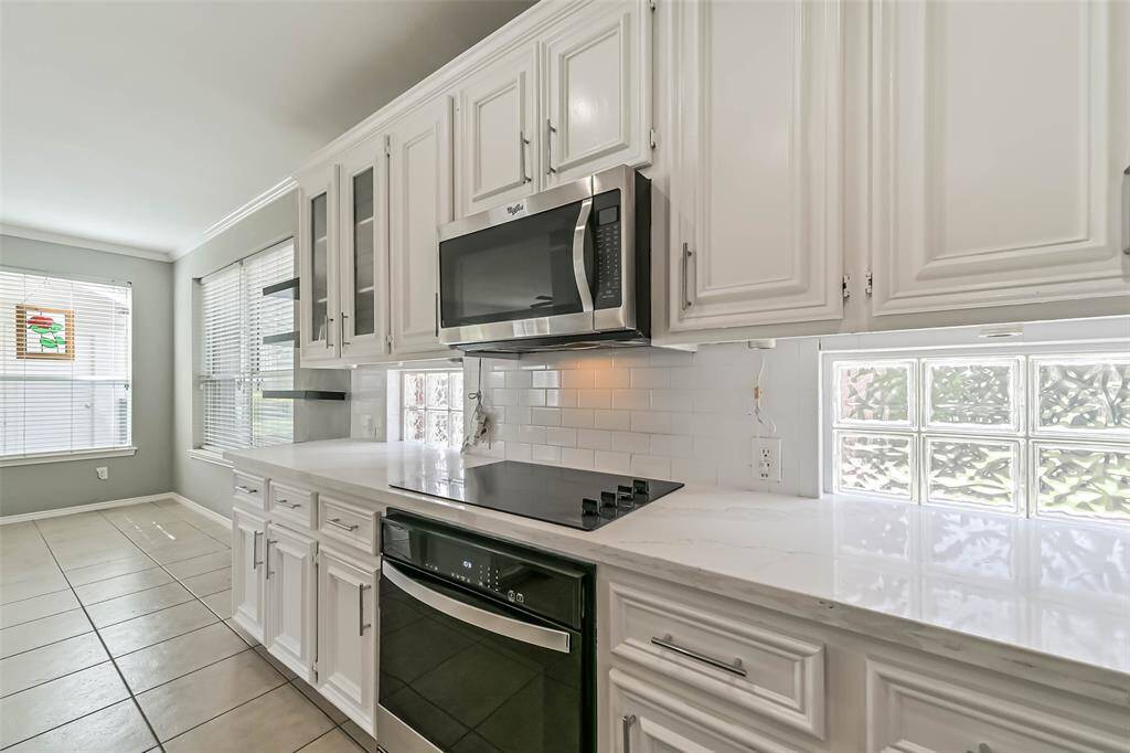 This bright kitchen features modern white cabinetry, a sleek cooktop, and stainless steel appliances. Large windows and glass blocks provide ample natural light, enhancing the spacious feel.