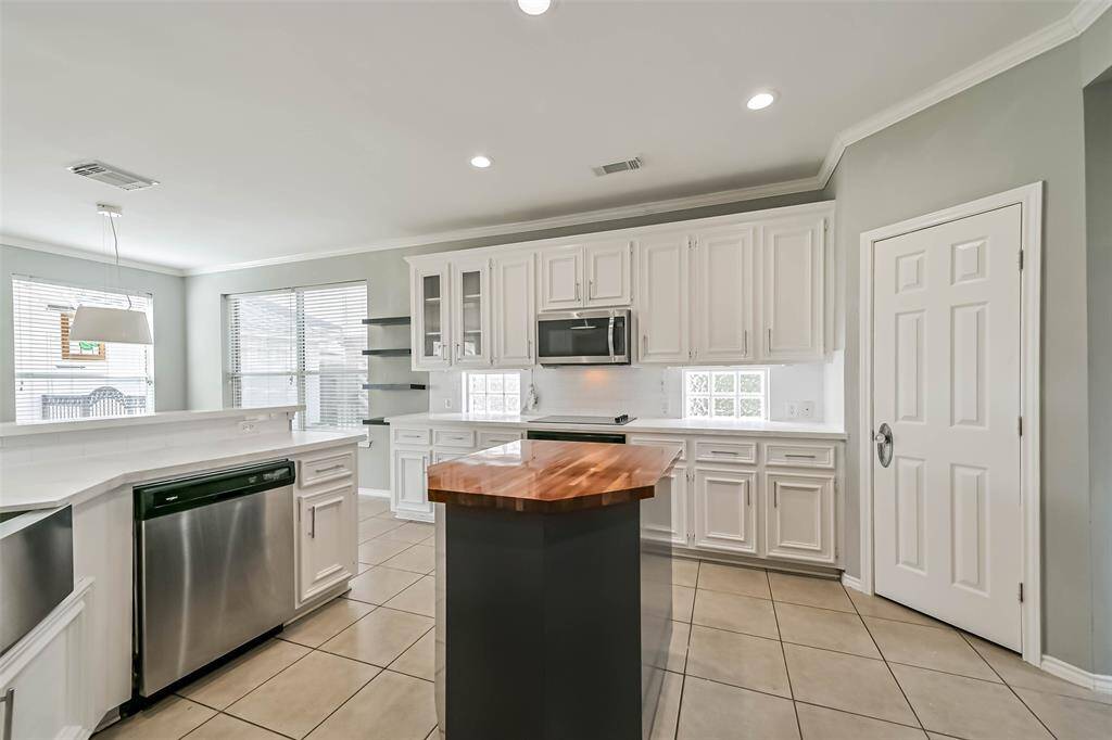This kitchen features modern stainless steel appliances, white cabinetry, and a center island with a wooden countertop. It has ample natural light from large windows and a spacious, open layout with tiled flooring.