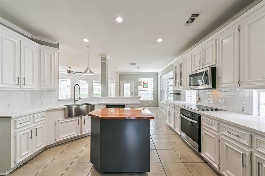 This spacious, modern kitchen features white cabinetry, stainless steel appliances, and a central island with a wooden countertop. The open layout provides ample natural light, and the sleek design includes a farmhouse sink and tiled flooring.