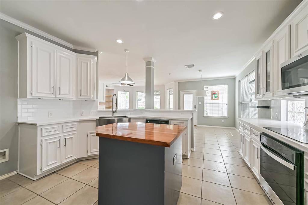 This kitchen features a modern design with white cabinetry, a central island with a wooden countertop, and stainless steel appliances. The space is bright and open, with recessed lighting and large windows providing plenty of natural light.