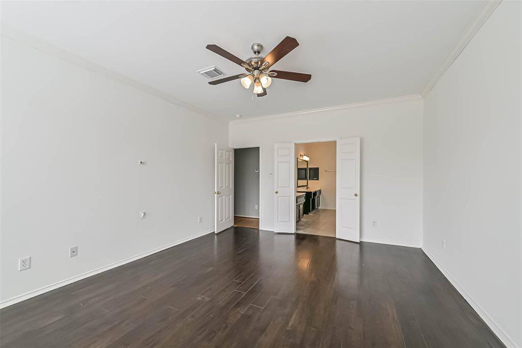 Spacious bedroom with dark hardwood floors, neutral walls, and a ceiling fan. It features double doors leading to a bathroom with dual sinks. Bright and modern.