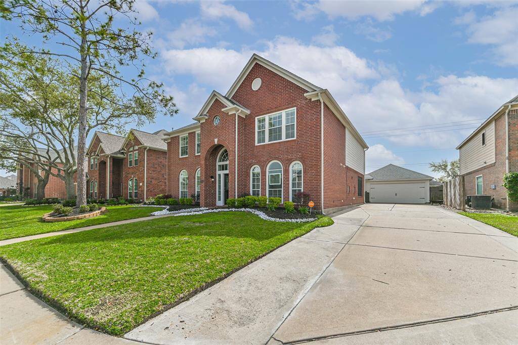 This photo showcases a spacious, two-story brick home with an elegant arched entryway and large windows. It features a well-maintained front lawn with mature trees and a long driveway leading to a detached garage. The neighborhood appears to be clean and well-kept.