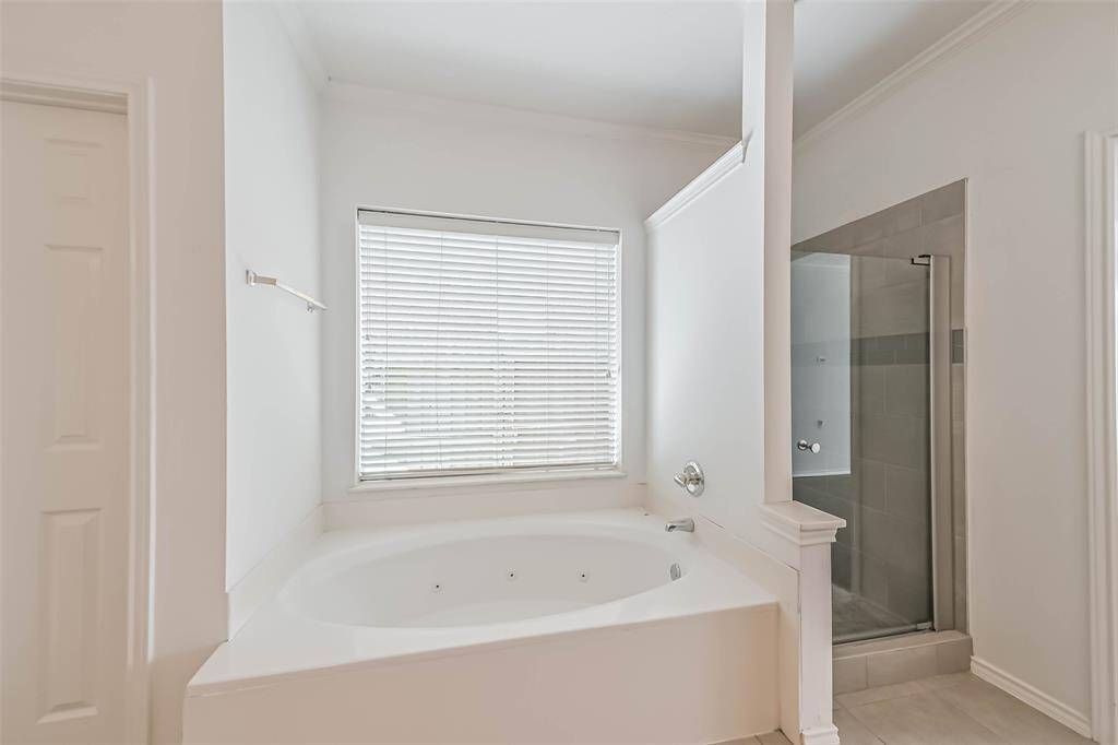 This bathroom features a spacious corner bathtub beneath a large window with blinds, offering plenty of natural light. Adjacent is a sleek, glass-enclosed shower, all set in a clean, bright space.