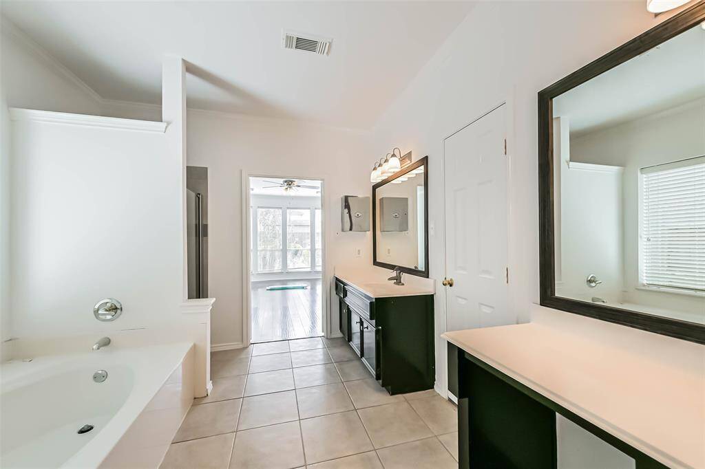 Bright, spacious bathroom with dual sinks, large mirrors, a soaking tub, and tile flooring. It features a doorway leading to a sunlit room, enhancing the open feel.