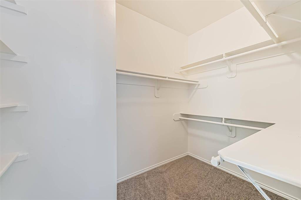 This photo shows a spacious walk-in closet with white walls, carpeted flooring, and multiple shelves and hanging rods for ample storage.