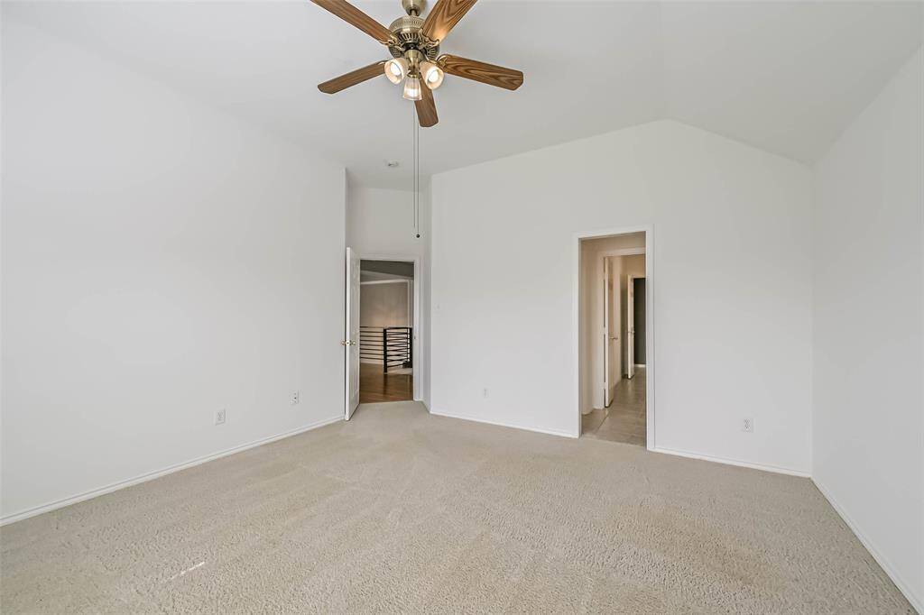 This photo shows a spacious, carpeted room with high ceilings and a ceiling fan. It features neutral walls, two doorways leading to a hallway and another room, and ample natural light.