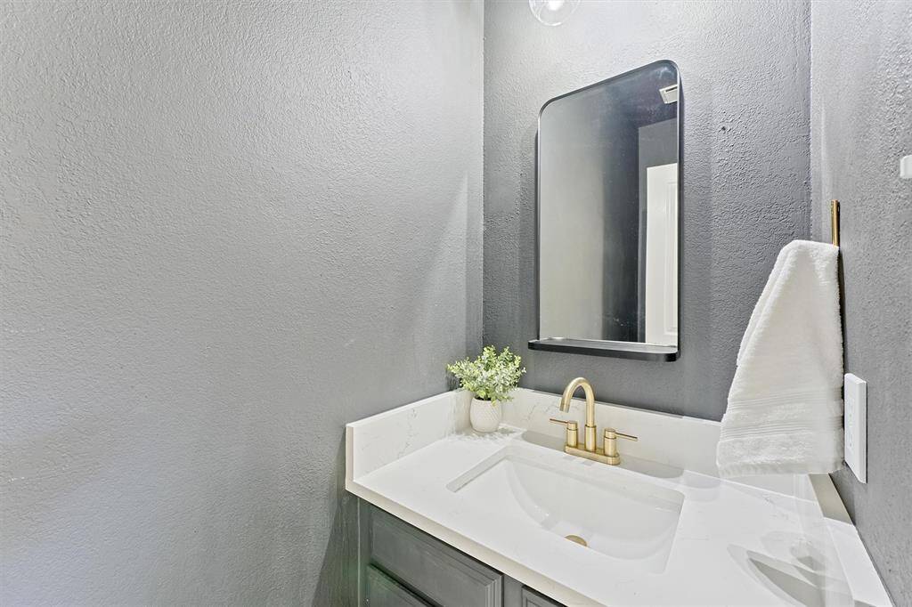 The half bathroom is modern, with a sleek, contemporary aesthetic. A white countertop and a gold faucet stand out against a calming backdrop of grey walls. The greenery gives the bathroom a refreshed vibe, while the contemporary mirror enhances the beauty of the space.