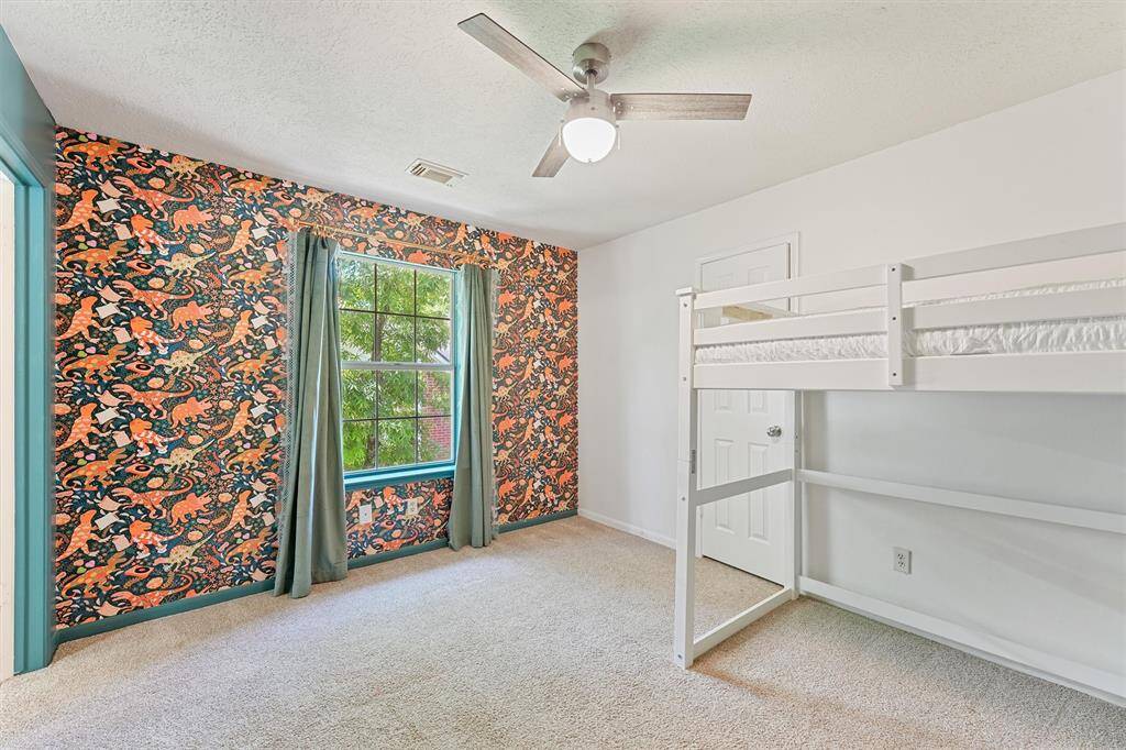 This is a bright bedroom that has an accent wall decorated with playful animal wallpaper. This light-filled bedroom includes a ceiling fan and carpeted flooring. A large window with green curtains adds to the sunny disposition of this cozy bedroom.