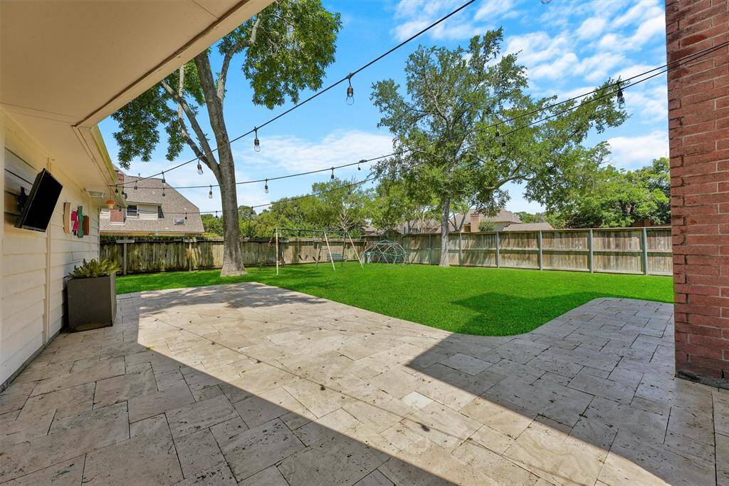 The expansive backyard features a large, paved patio area perfect for outdoor gatherings, surrounded by a lush green lawn and mature trees. String lights add ambiance, and a wooden fence provides privacy. Ideal for entertaining and relaxation.