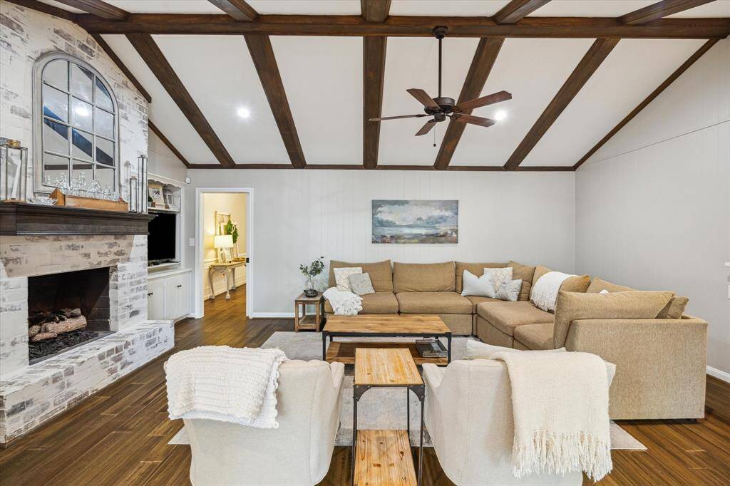 This inviting living room features a cozy sectional sofa, wooden ceiling beams, and a rustic brick fireplace. It has a warm, neutral palette and plenty of natural light, creating a comfortable and spacious atmosphere.